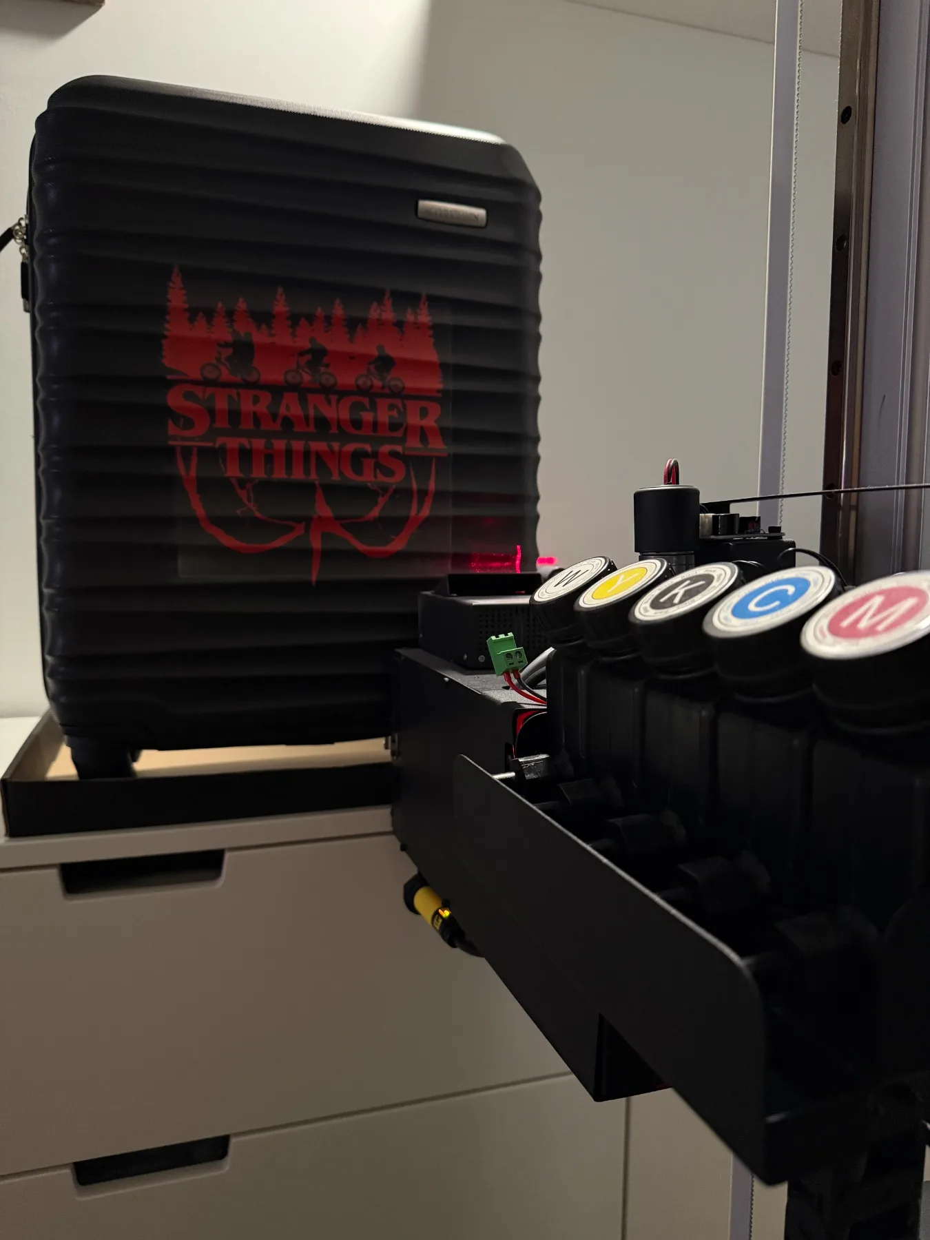 Czerwona grafika Stranger Things wydrukowana na czarnej walizce, obok głowica drukarki z tuszami CMYK